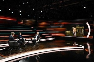 Personen diskutieren in einem TV-Studio mit zugeschaltetem Interviewpartner auf einem Bildschirm.